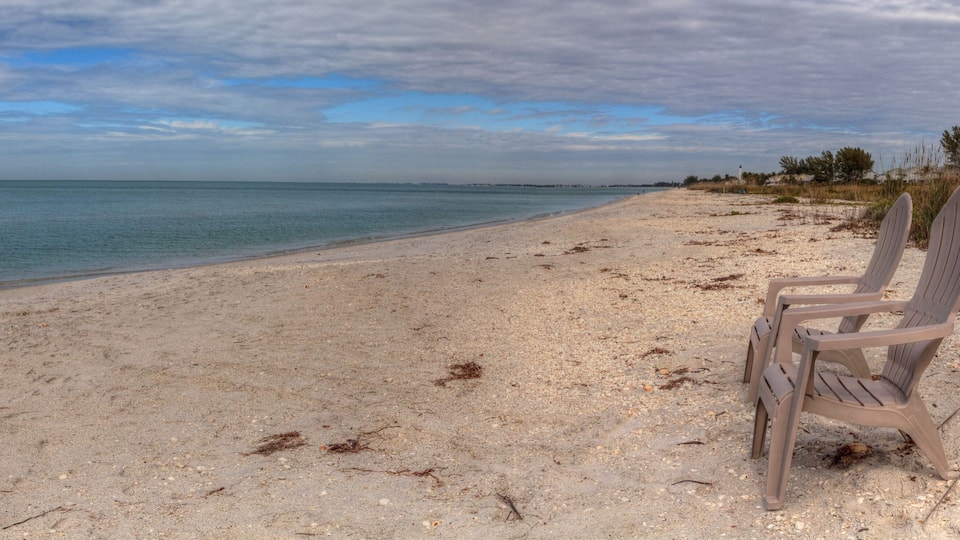 A beach with a single plastic chair, clear blue water, and a cloudy sky.