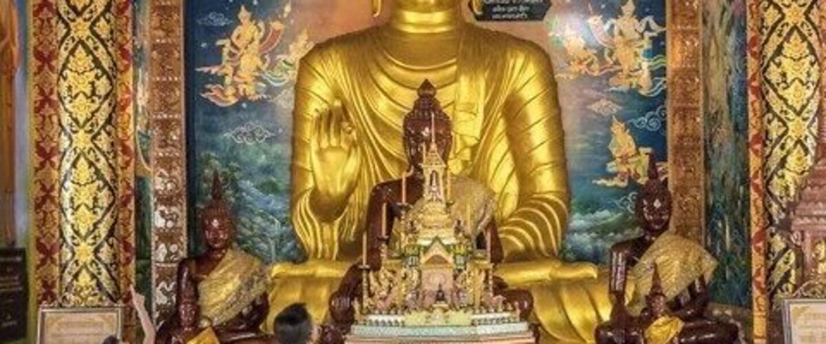 A large golden Buddha statue in a temple with intricate wall paintings and ornate decorations.