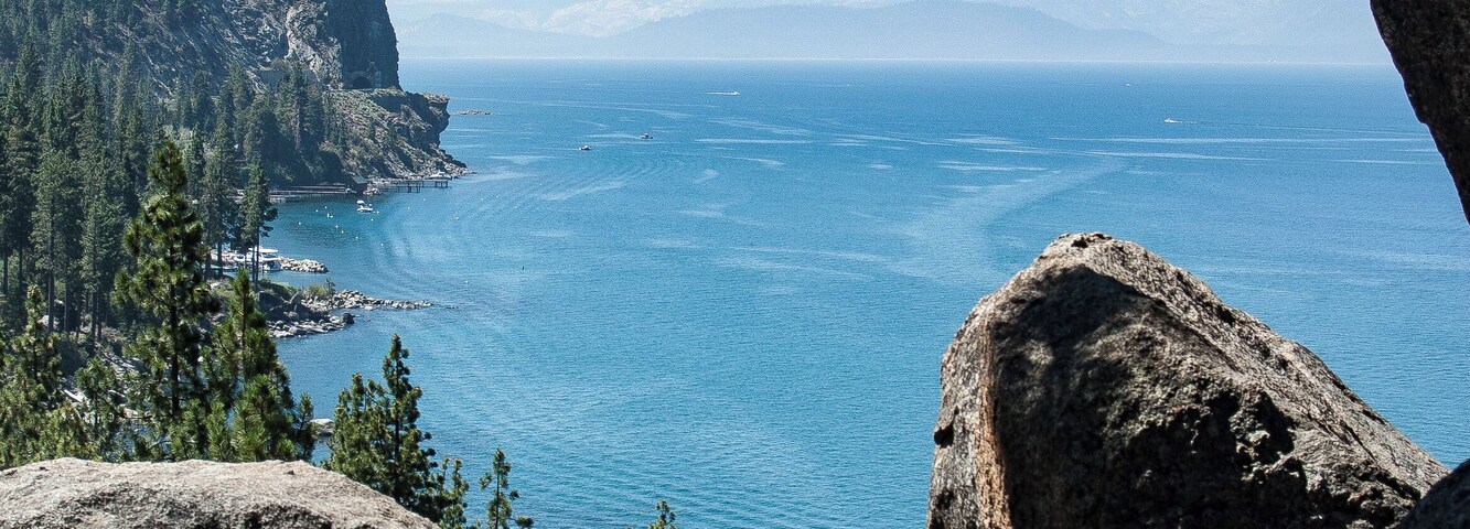 Paisagem costeira com grandes pedras, densos pinheiros e mar azul cristalino.
