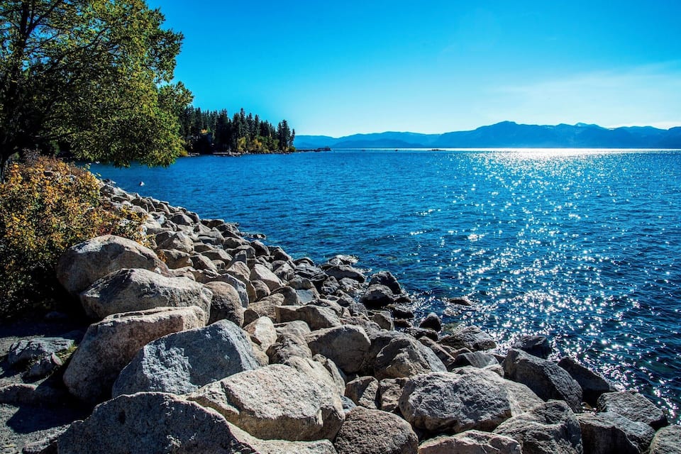 A rocky shoreline with a calm lake, trees, and distant mountains.