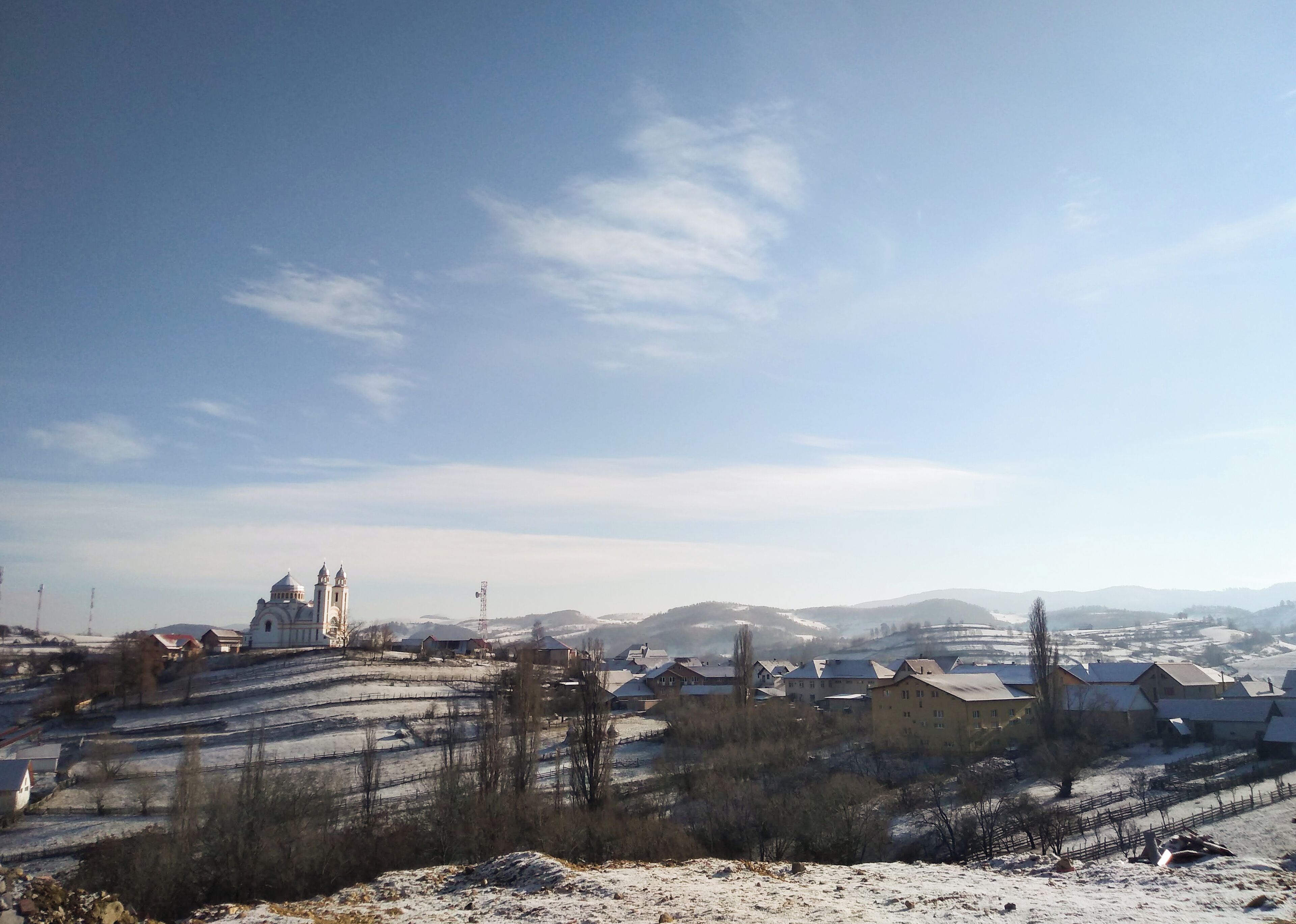 A snow-covered landscape with a church, residential buildings, and rolling hills in the background.