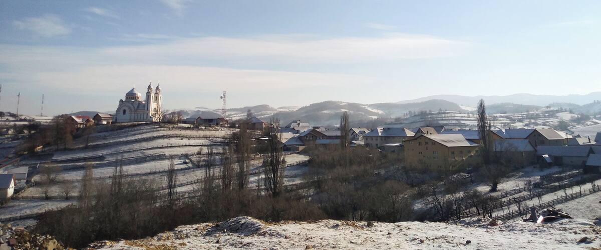 A snow-covered landscape with a church, residential buildings, and rolling hills in the background.