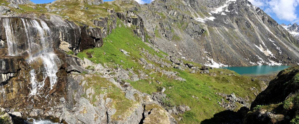 A mountainous landscape with a waterfall, green grass, rocky terrain, and a turquoise lake.