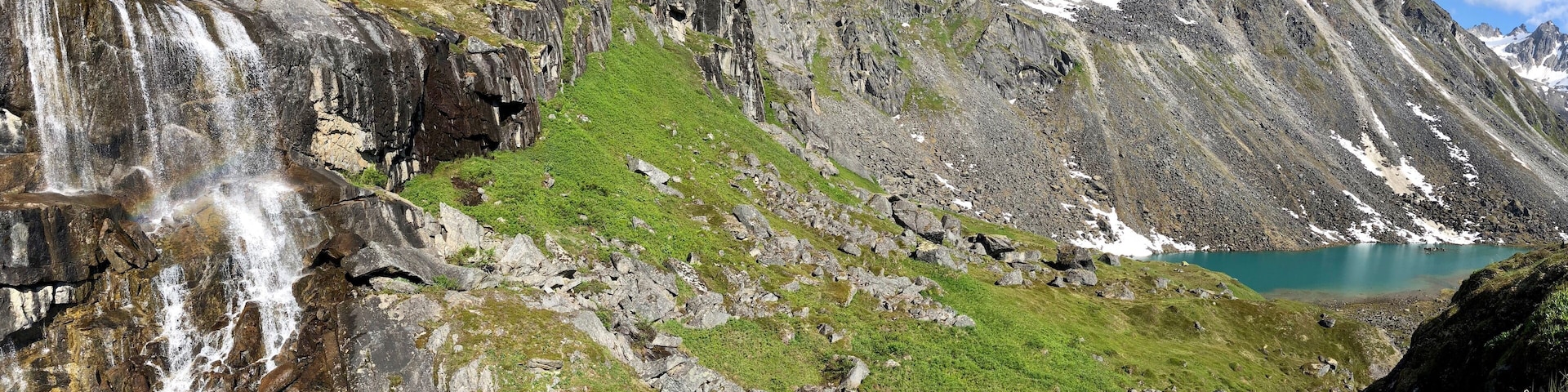 A mountainous landscape with a waterfall, green grass, rocky terrain, and a turquoise lake.