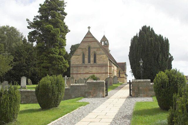 A church with a pointed roof and a cross on top, surrounded by a cemetery with headstones and trimmed bushes.