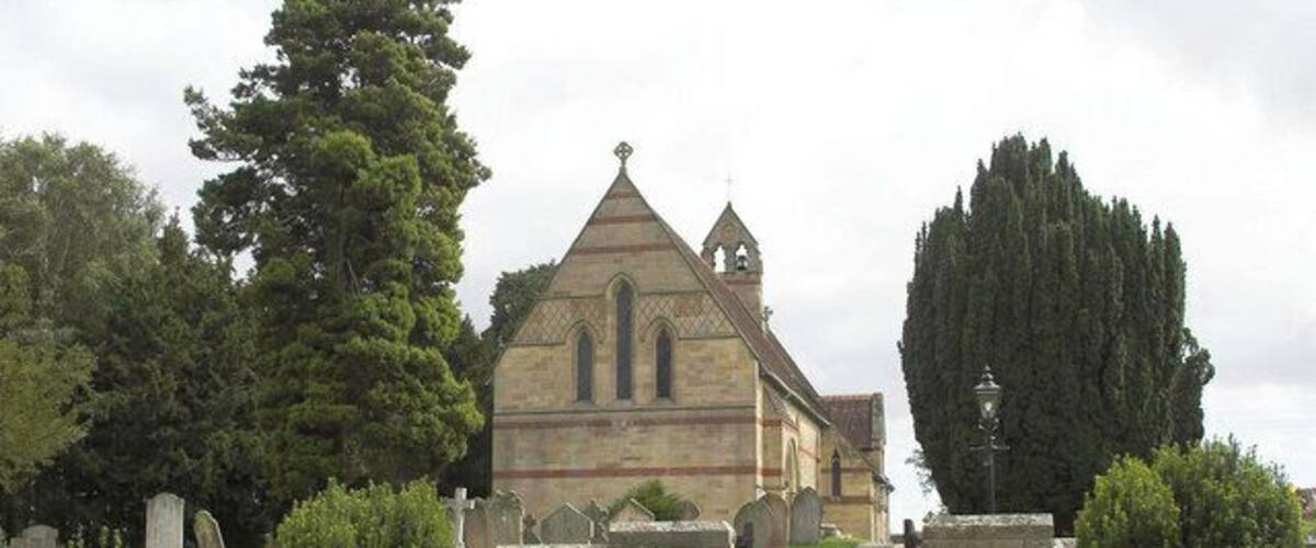 A church with a pointed roof and a cross on top, surrounded by a cemetery with headstones and trimmed bushes.