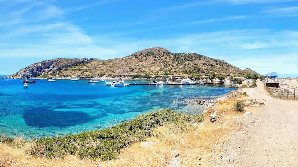 A coastal scene with clear blue waters, boats, a rocky hill, and a dirt path.