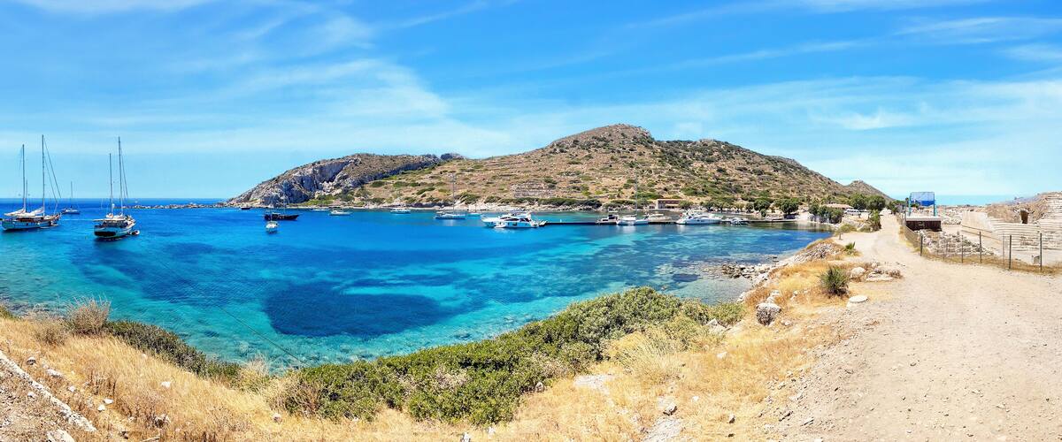 A coastal scene with clear blue waters, boats, a rocky hill, and a dirt path.