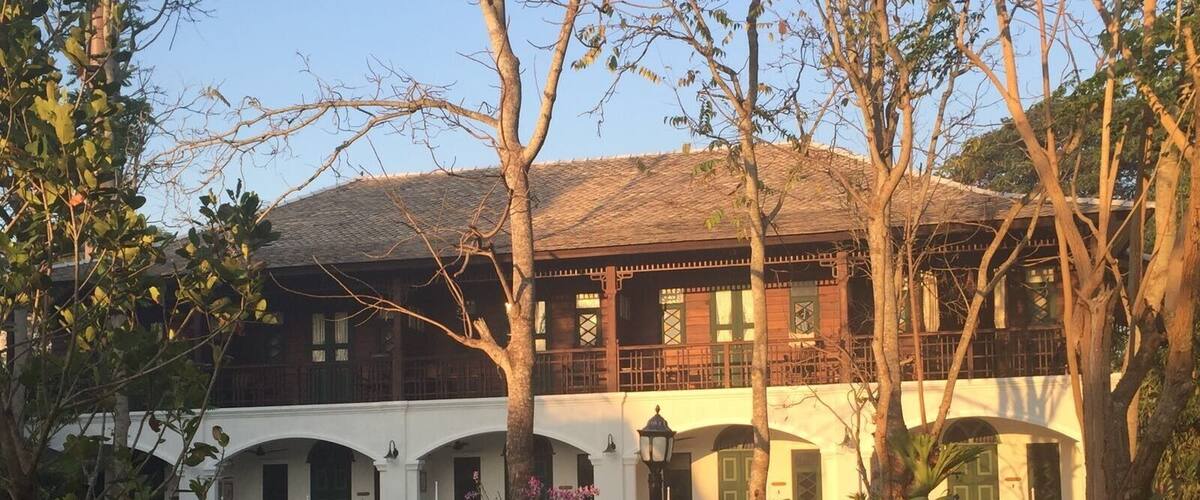 A traditional building with a tiled roof, white walls, and green doors. There are trees and a bench in the courtyard.