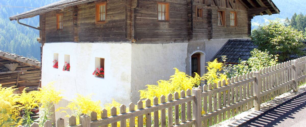 A wooden house with a steep roof, surrounded by a wooden fence and yellow flowering plants.