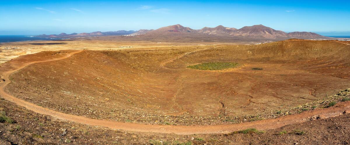 A barren landscape with a large crater, a winding dirt road, and distant mountains.