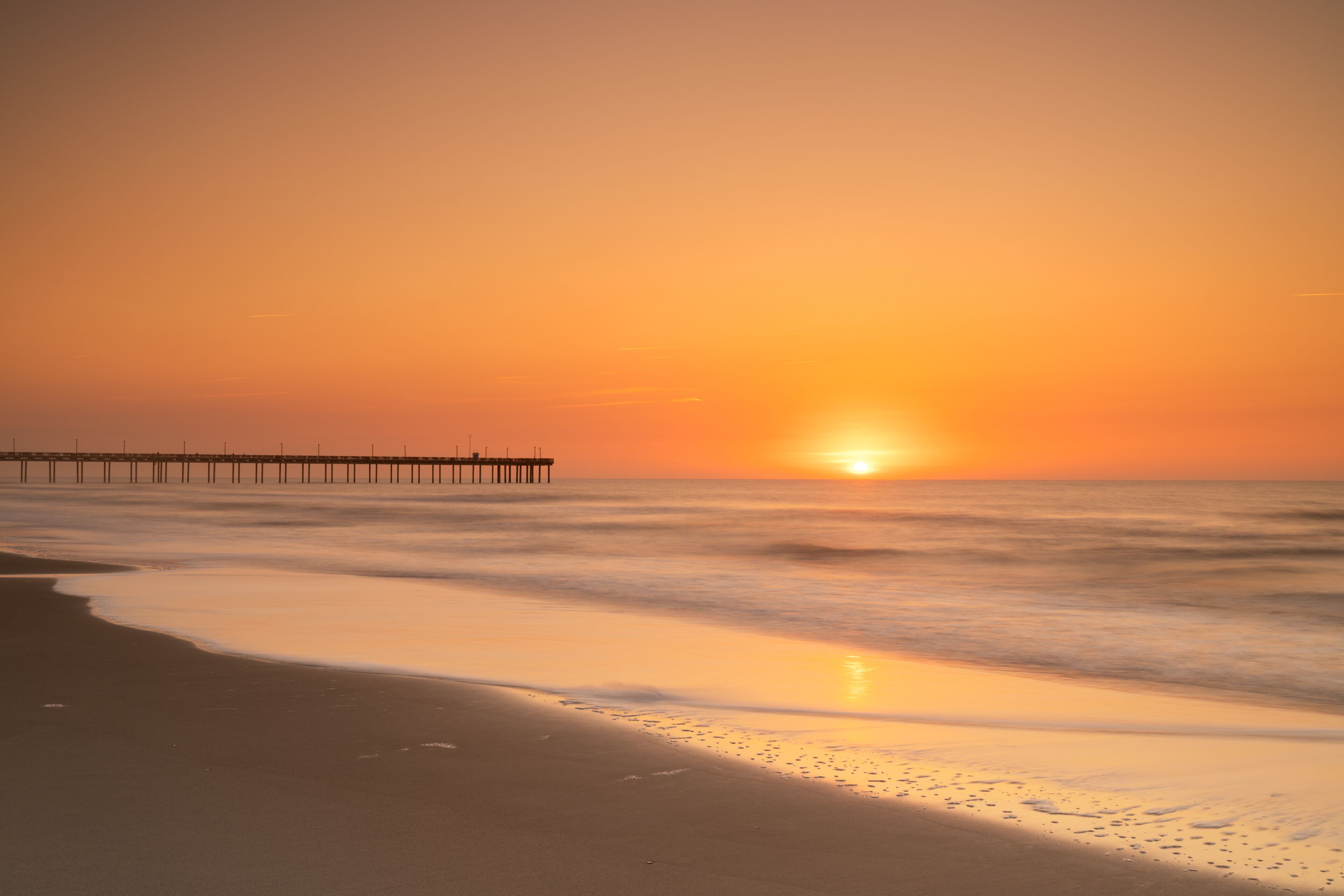 A sunset over a beach with waves and a pier.