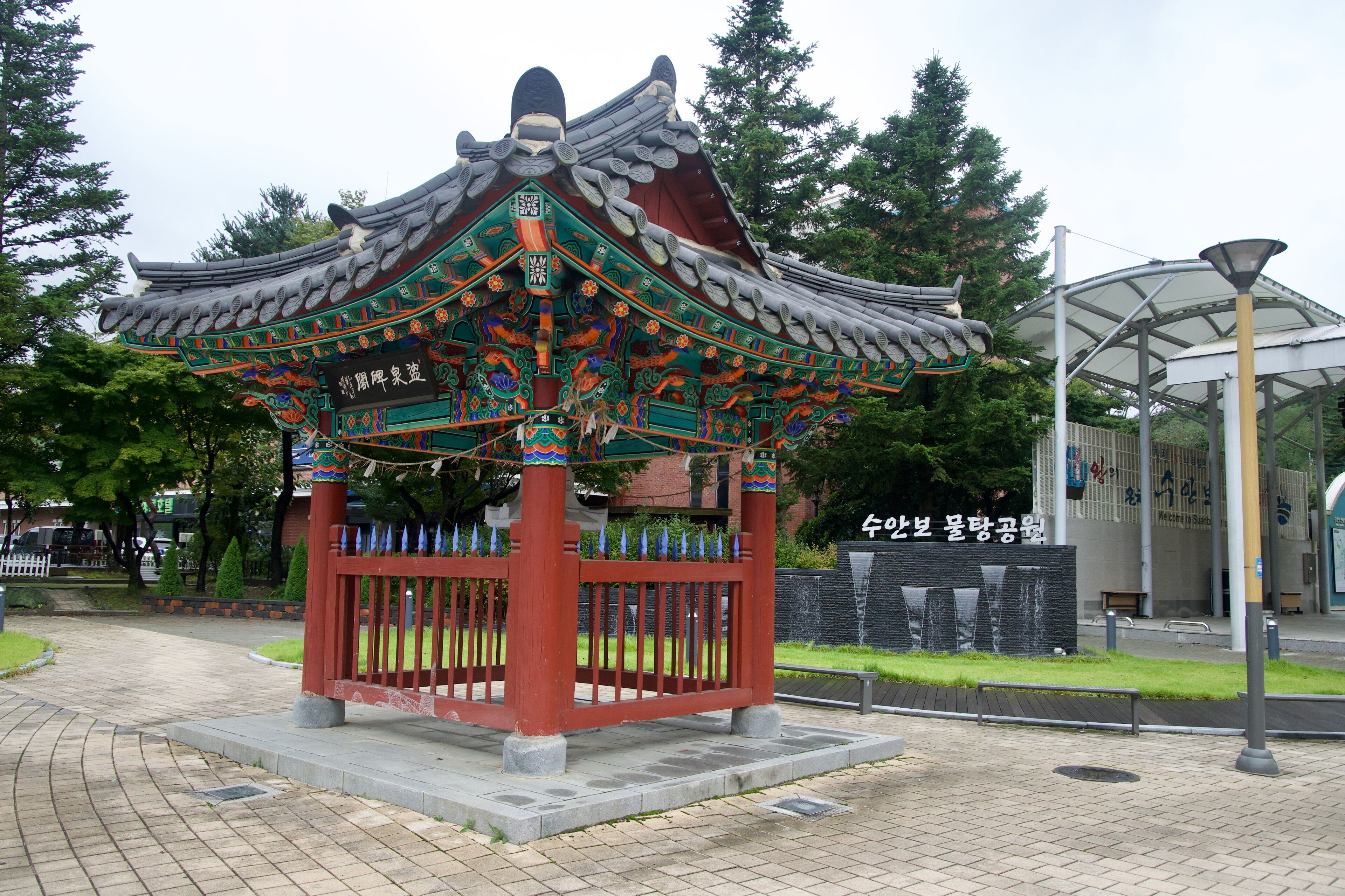 A traditional Korean pavilion with ornate roof and colorful decorations, surrounded by a paved walkway and greenery.