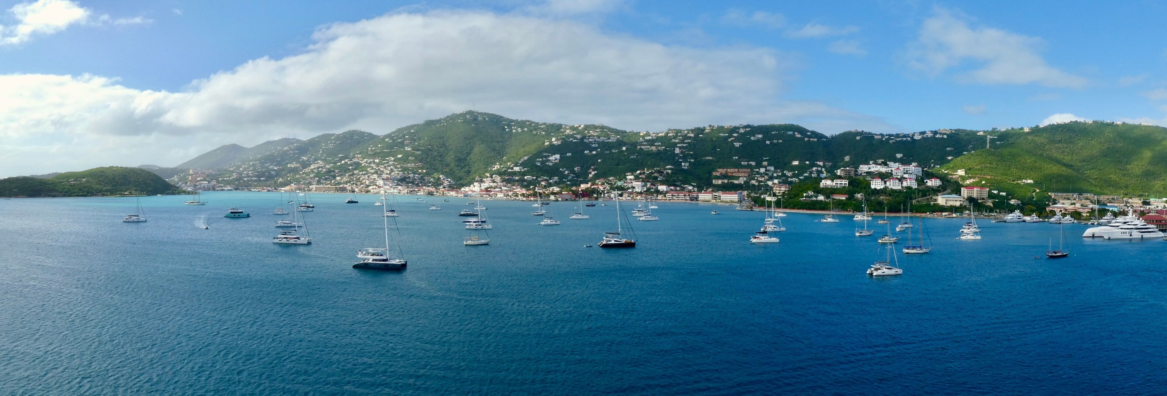 A coastal town with numerous sailboats anchored in the bay, surrounded by lush green hills and a clear blue sky.