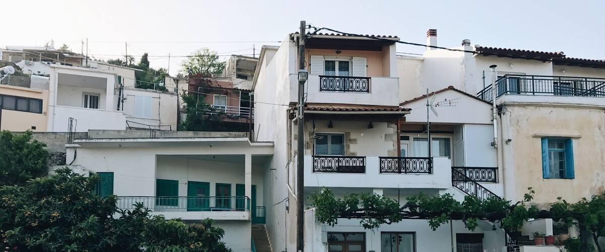 A row of white buildings with balconies and green shutters, surrounded by trees and potted plants.