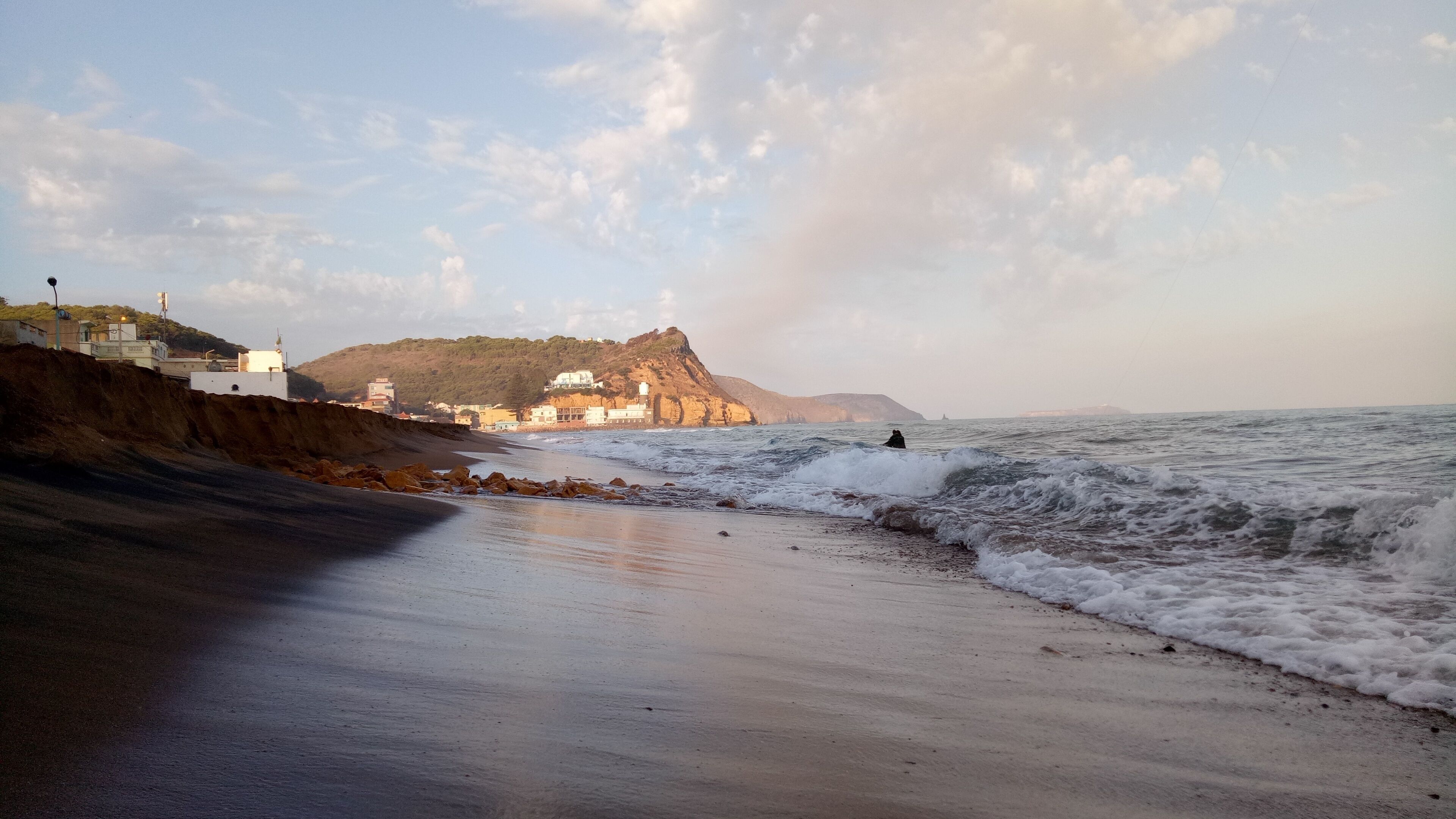 A beach with waves, a cliff with buildings, and a person surfing.