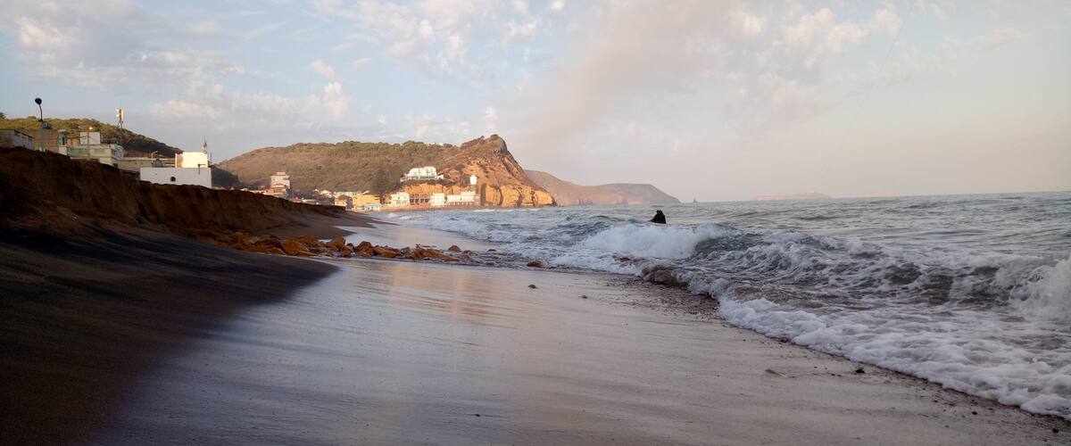 A beach with waves, a cliff with buildings, and a person surfing.
