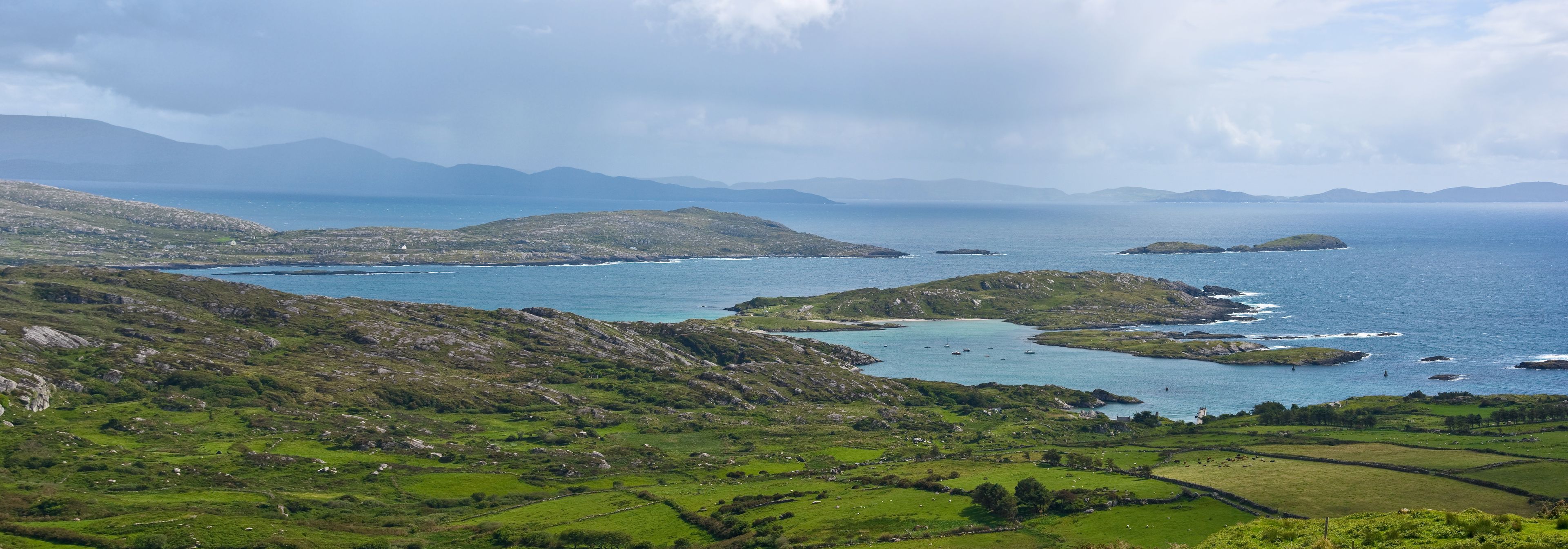 Derrynane Bay, Kerry, Ireland. Rocky islands in a blue sea with distant mountains visible through rain showers, and a rugged pasture of green fields and stone walls in the foreground.