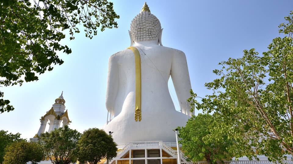 A large white Buddha statue with a golden sash, surrounded by trees and a smaller temple in the background.
