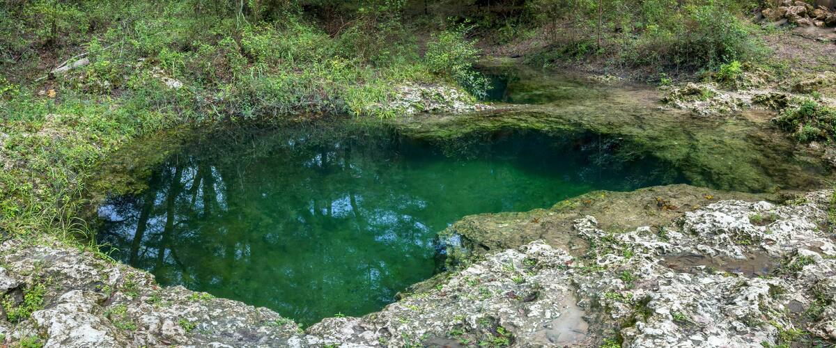 A clear, circular natural pool surrounded by rocky terrain and dense greenery.