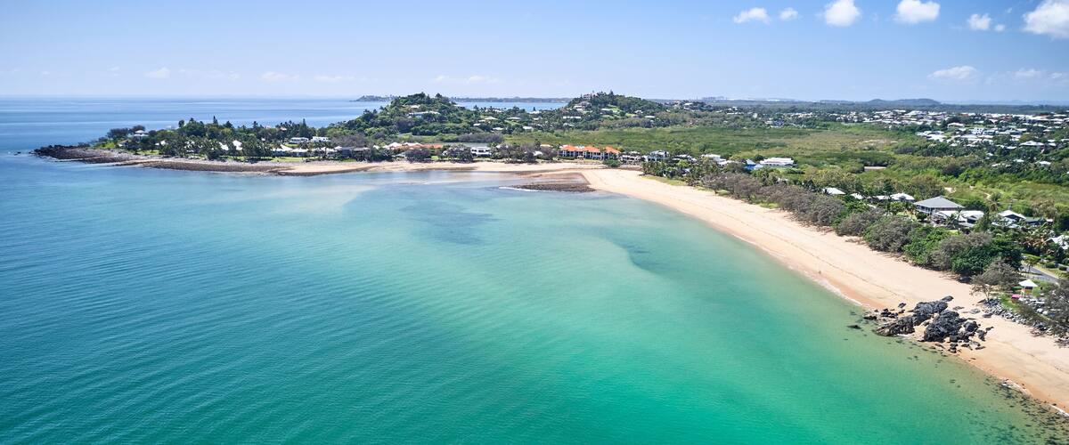 A coastal landscape with a sandy beach, clear turquoise waters, and a hilly background with scattered buildings.