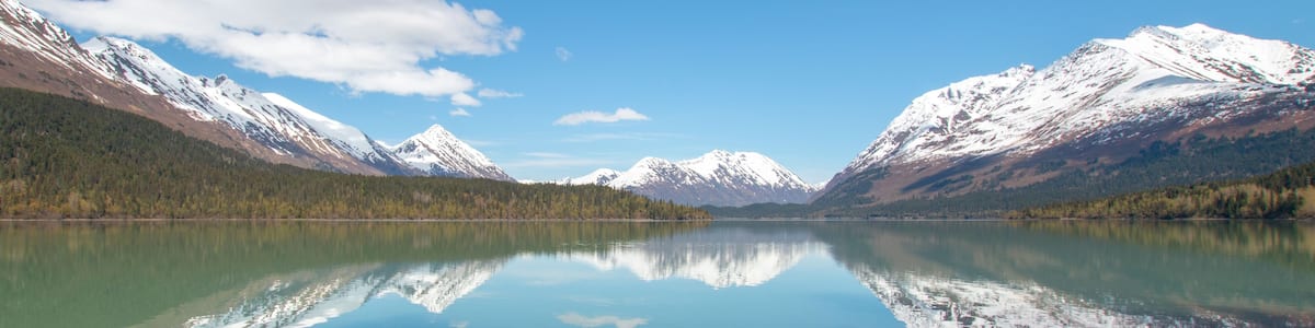 A calm lake reflecting snow-capped mountains, clear blue sky, and a forested valley.