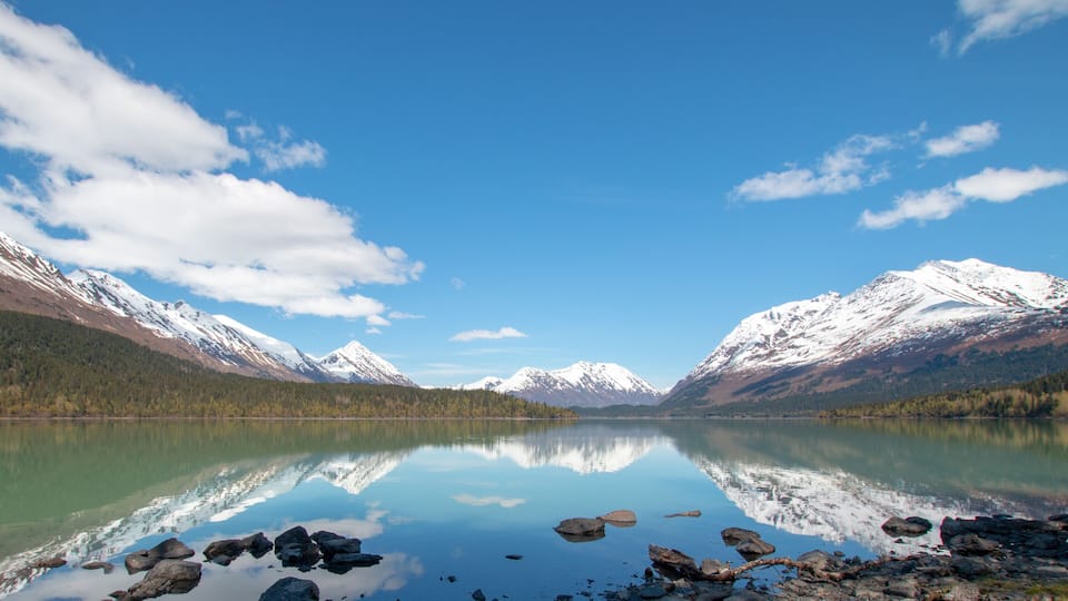A calm lake reflecting snow-capped mountains, clear blue sky, and a forested valley.