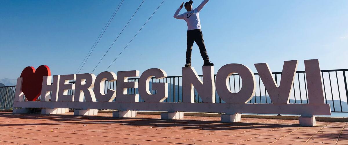 A person standing on a platform with a sign that reads "HERCEG NOVI".