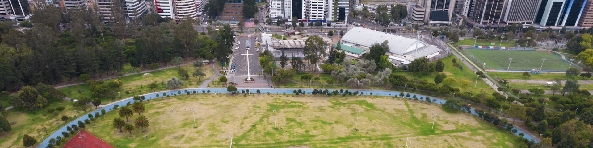 An aerial view of a large oval-shaped park with a running track, a red sports field, and a river running through the middle.