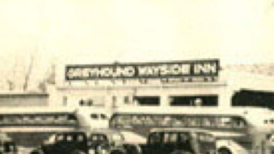 A vintage car in front of a building with a sign that reads "Greyhound Wayside Inn."