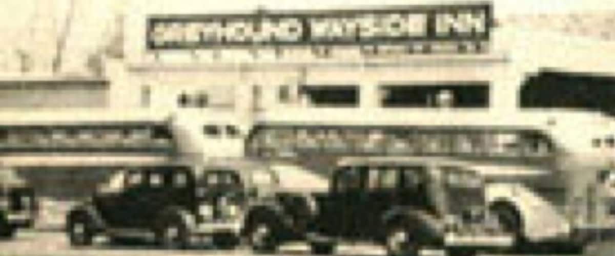 A vintage car in front of a building with a sign that reads "Greyhound Wayside Inn."