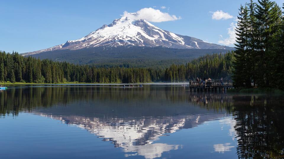 A calm lake with a reflection of a snow-capped mountain and dense forest.