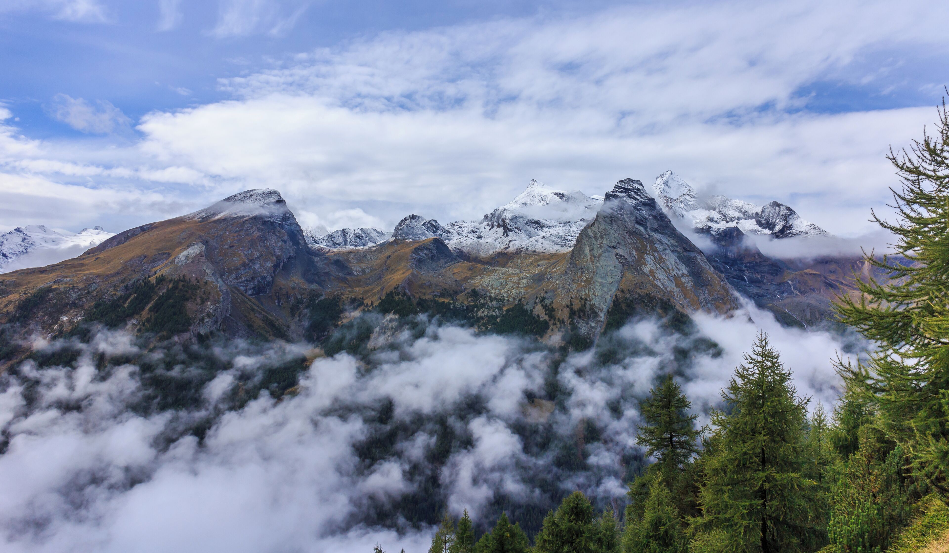 A mountain range with snow-capped peaks, dense forests, and clouds at the base.