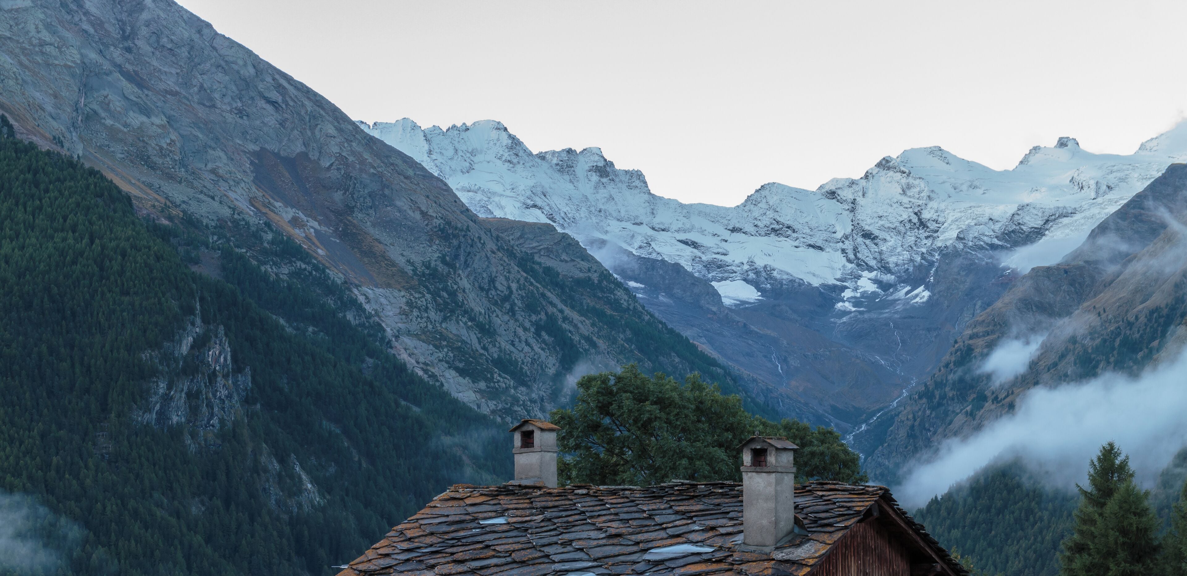 A mountain cabin with a stone roof, surrounded by snow-covered peaks and dense forest.
