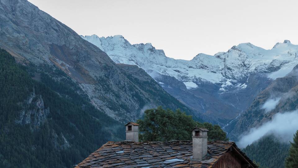 A mountain cabin with a stone roof, surrounded by snow-covered peaks and dense forest.