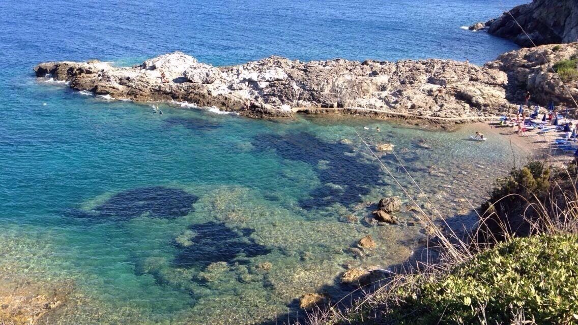 A rocky coastline with clear blue waters, a sandy beach, and a few people in the distance.