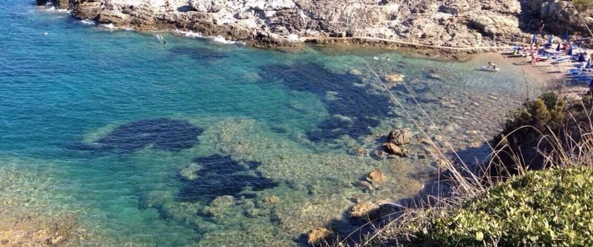 A rocky coastline with clear blue waters, a sandy beach, and a few people in the distance.