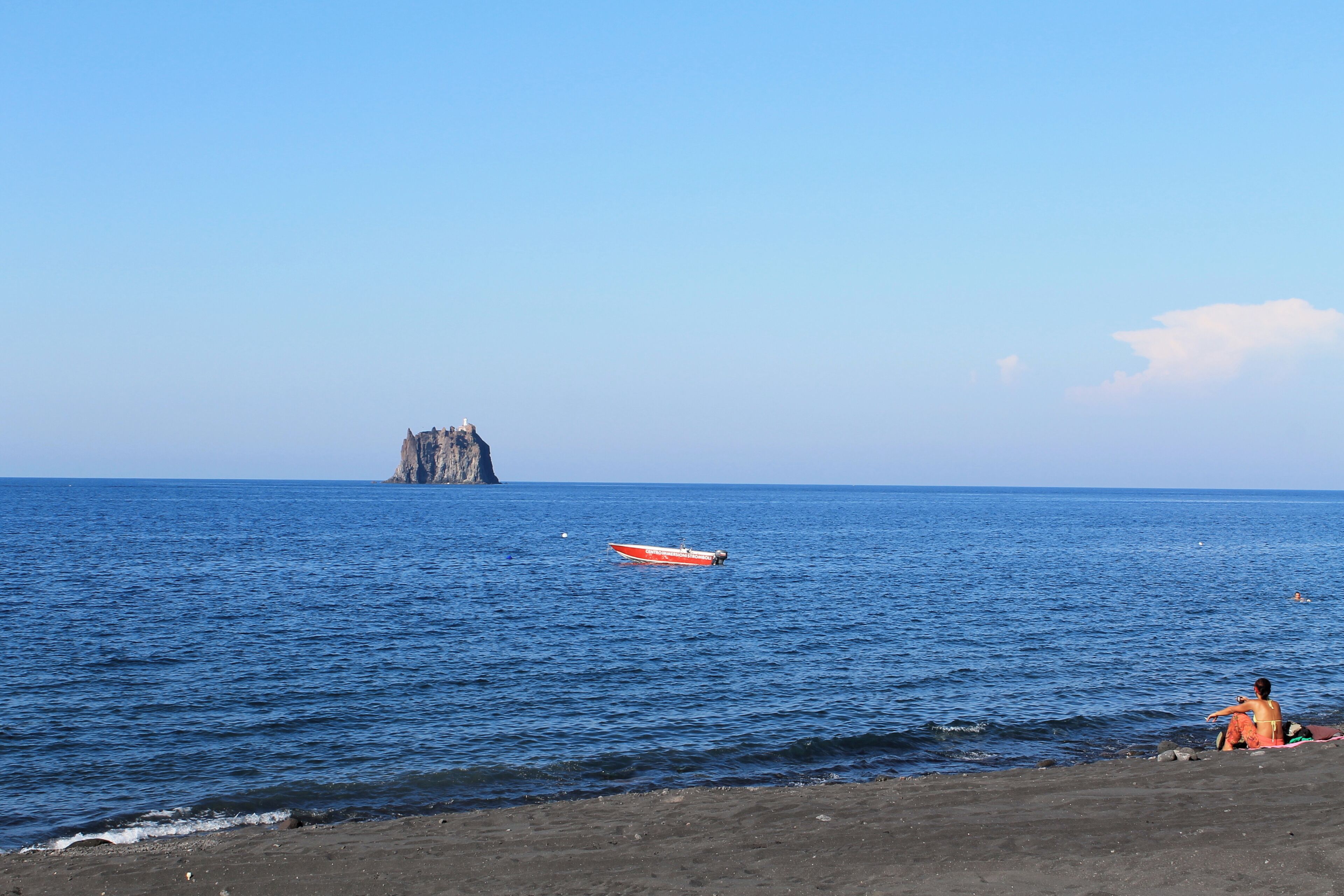 A beach with a rocky outcrop in the distance, a red boat on the water, and a person sitting on the sand.