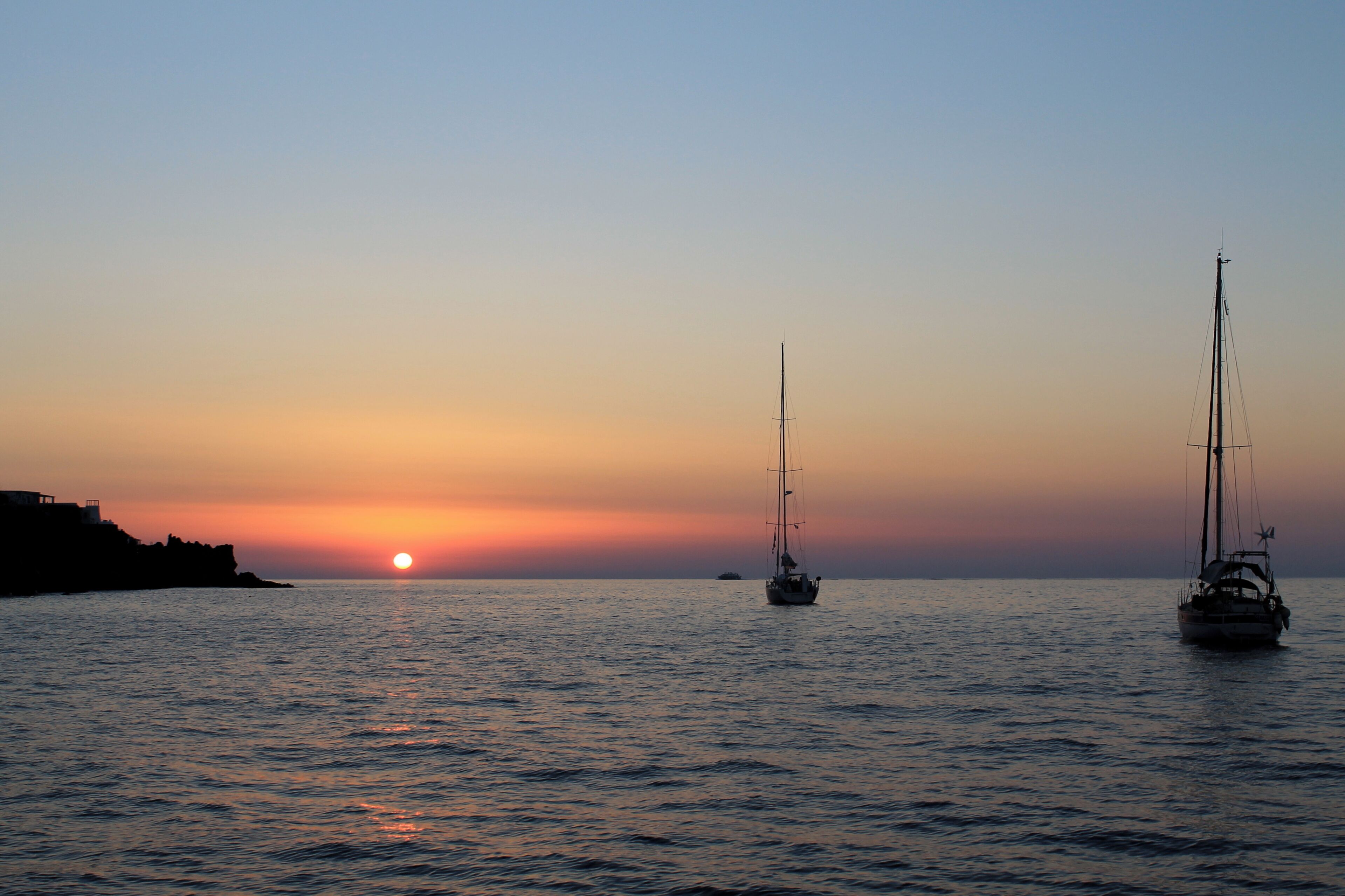 A sunset over calm waters with two sailboats.