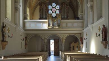 A church interior with a high arched ceiling, a chandelier, stained glass windows, and rows of pews.