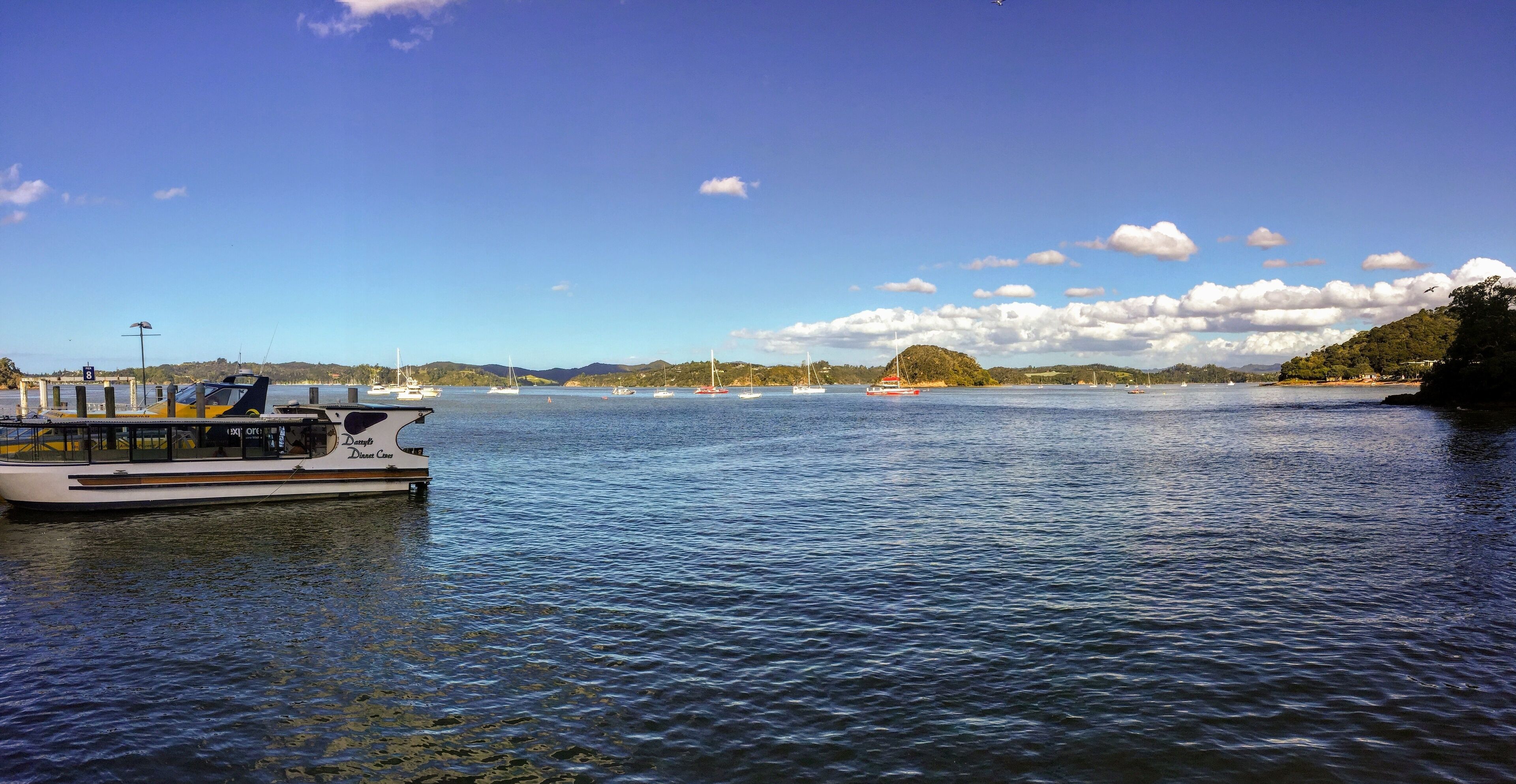 A calm lake with a boat docked on the left, sailboats in the distance, and a hilly landscape under a clear sky.