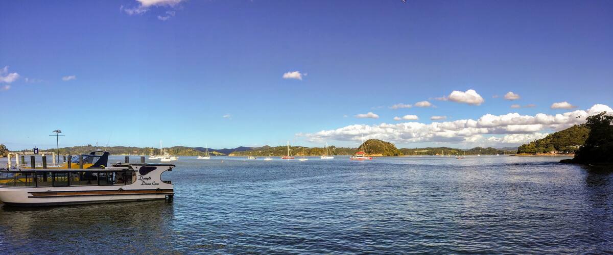 A calm lake with a boat docked on the left, sailboats in the distance, and a hilly landscape under a clear sky.