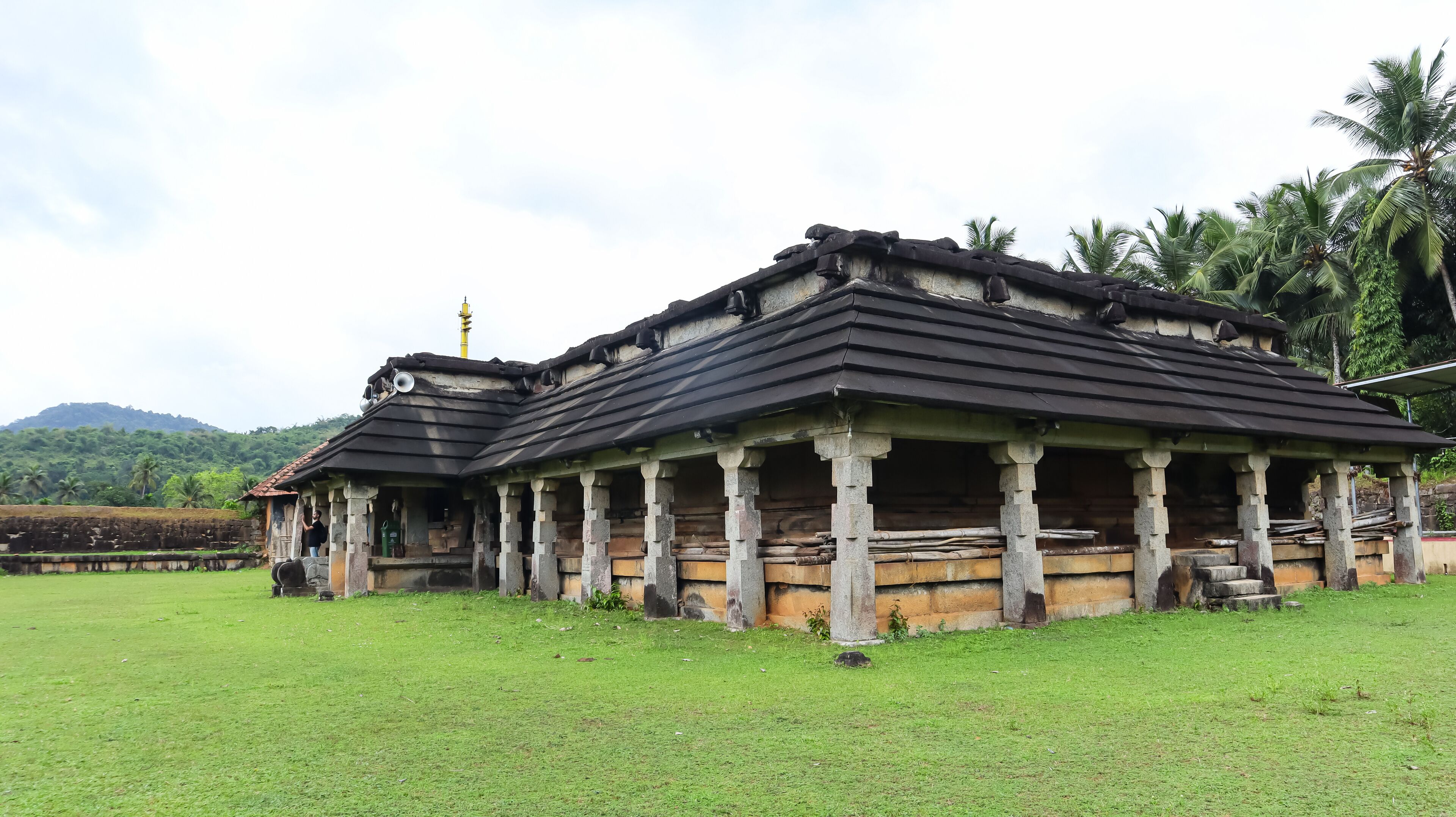 A traditional building with a tiled roof and stone pillars, surrounded by a grassy field and palm trees.