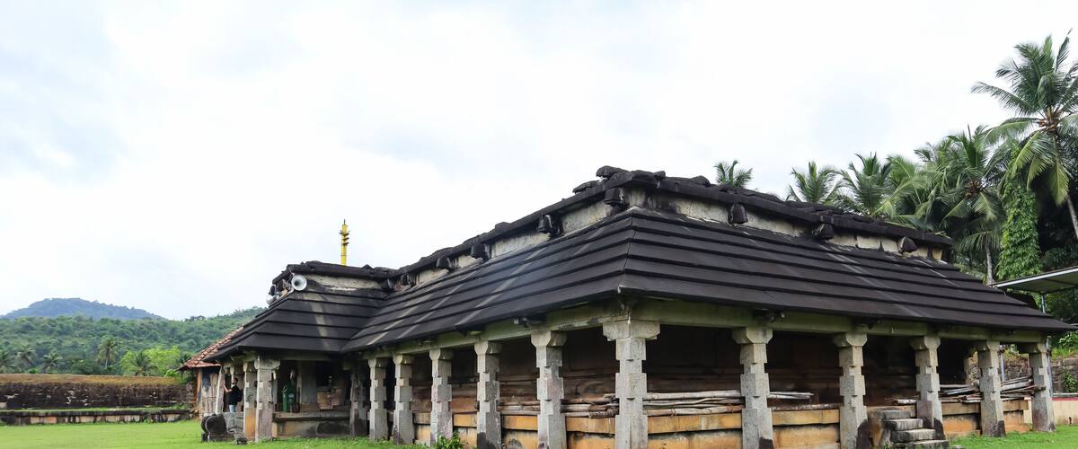 A traditional building with a tiled roof and stone pillars, surrounded by a grassy field and palm trees.