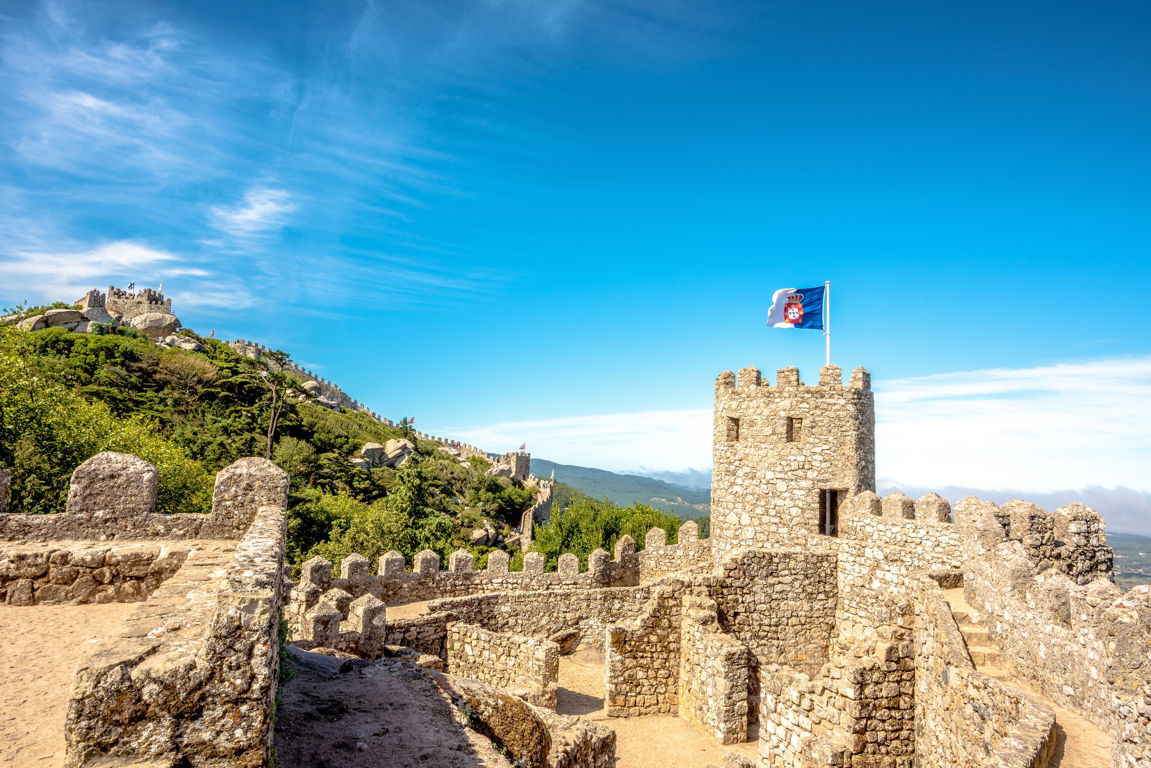 A historic stone fortress with a flag on top, surrounded by lush greenery and a clear blue sky.