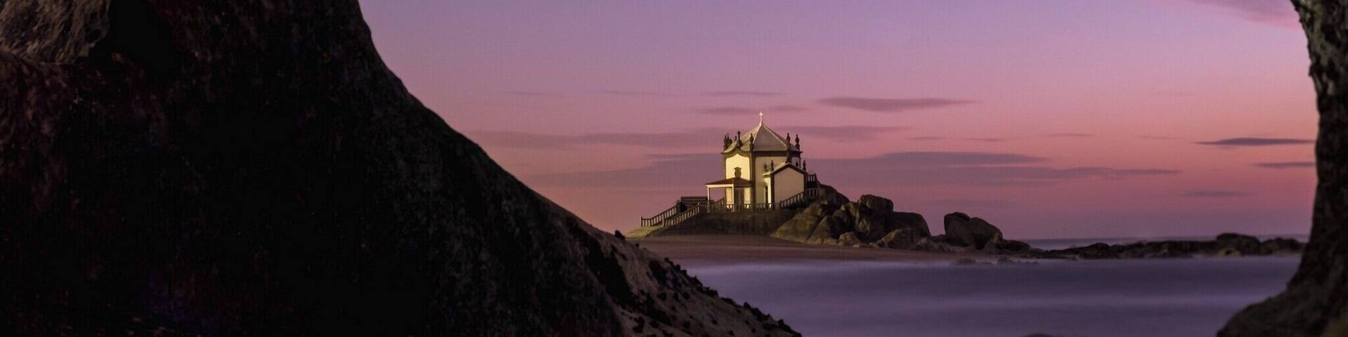 A coastal landscape with a small illuminated building on a rocky outcrop, surrounded by calm waters and dramatic rock formations.