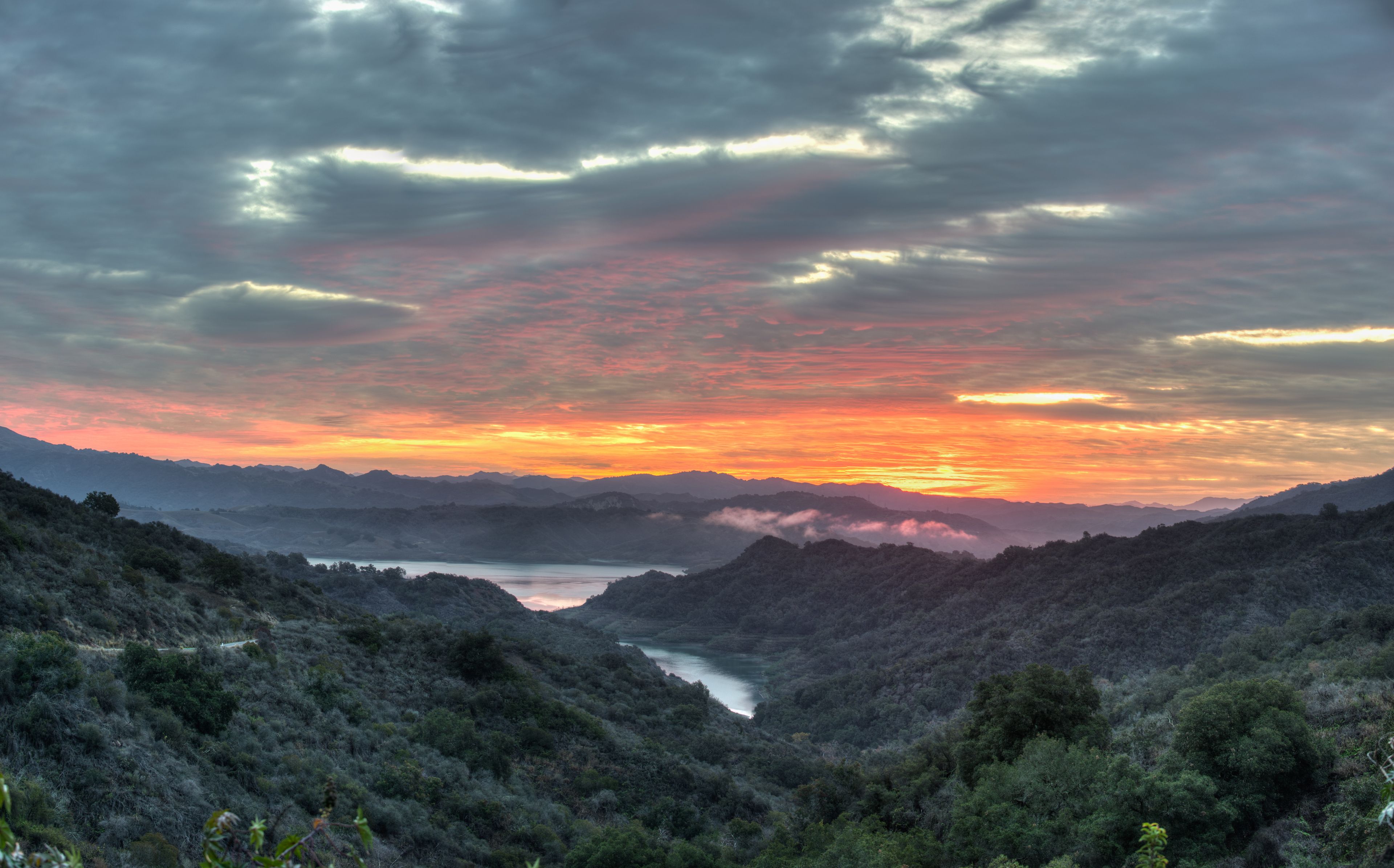 A mountainous landscape with a river valley, dense vegetation, and a sunset with clouds.