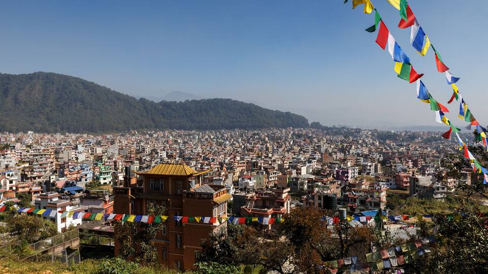 A cityscape with colorful prayer flags, a prominent building, and a mountainous backdrop.