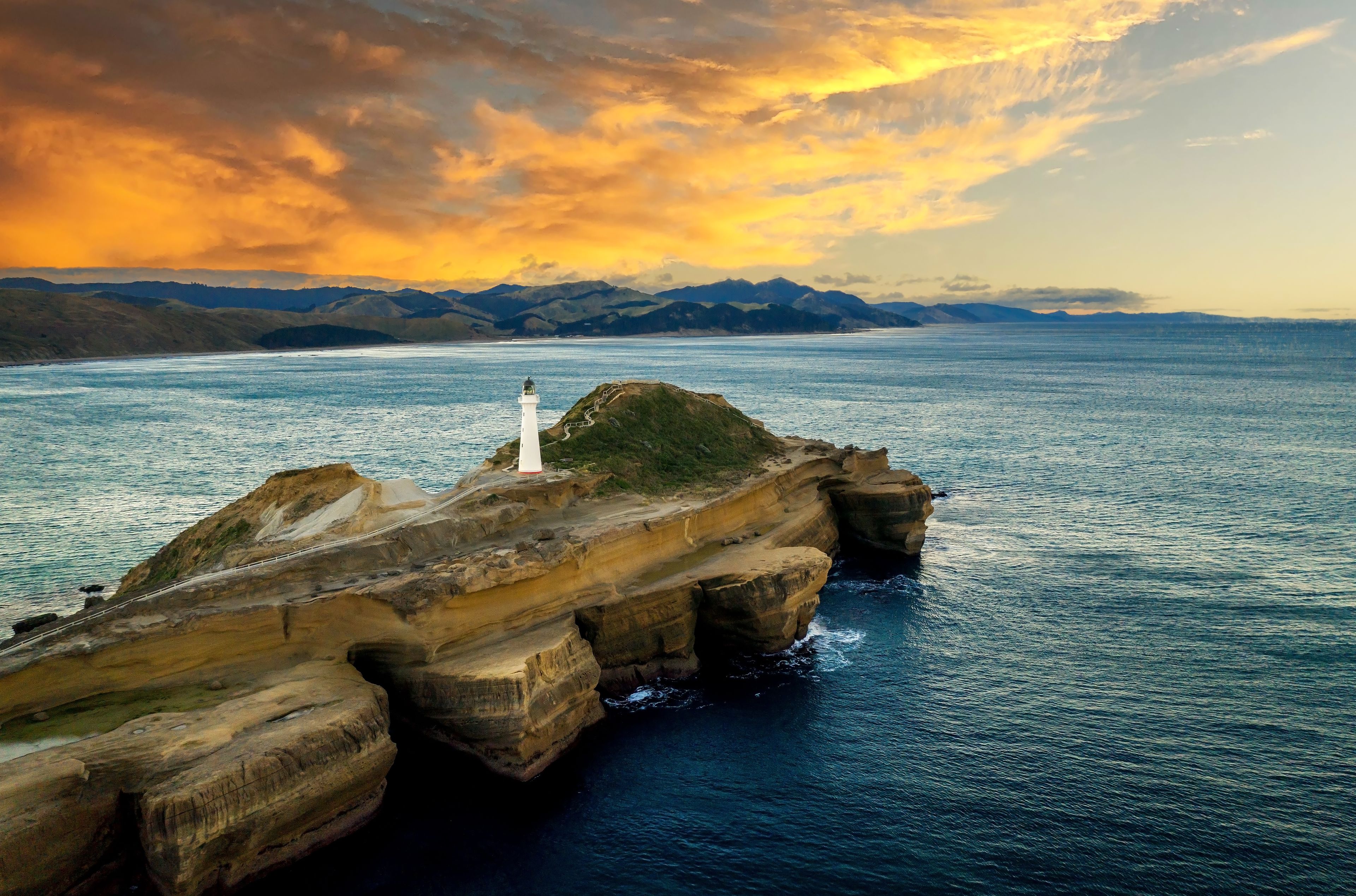 A coastal landscape with a lighthouse on a rocky outcrop, a clear sky, and distant mountains.