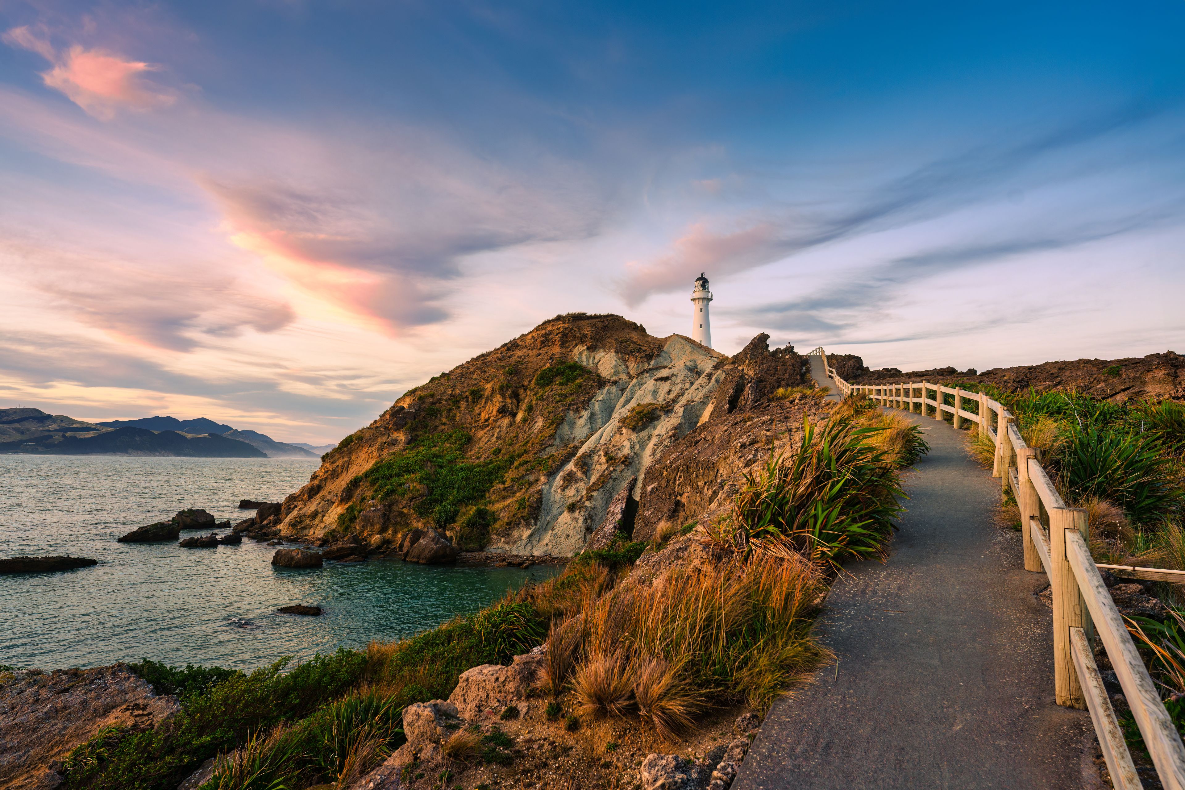A coastal landscape with a lighthouse on a rocky outcrop, a wooden walkway, and calm sea.